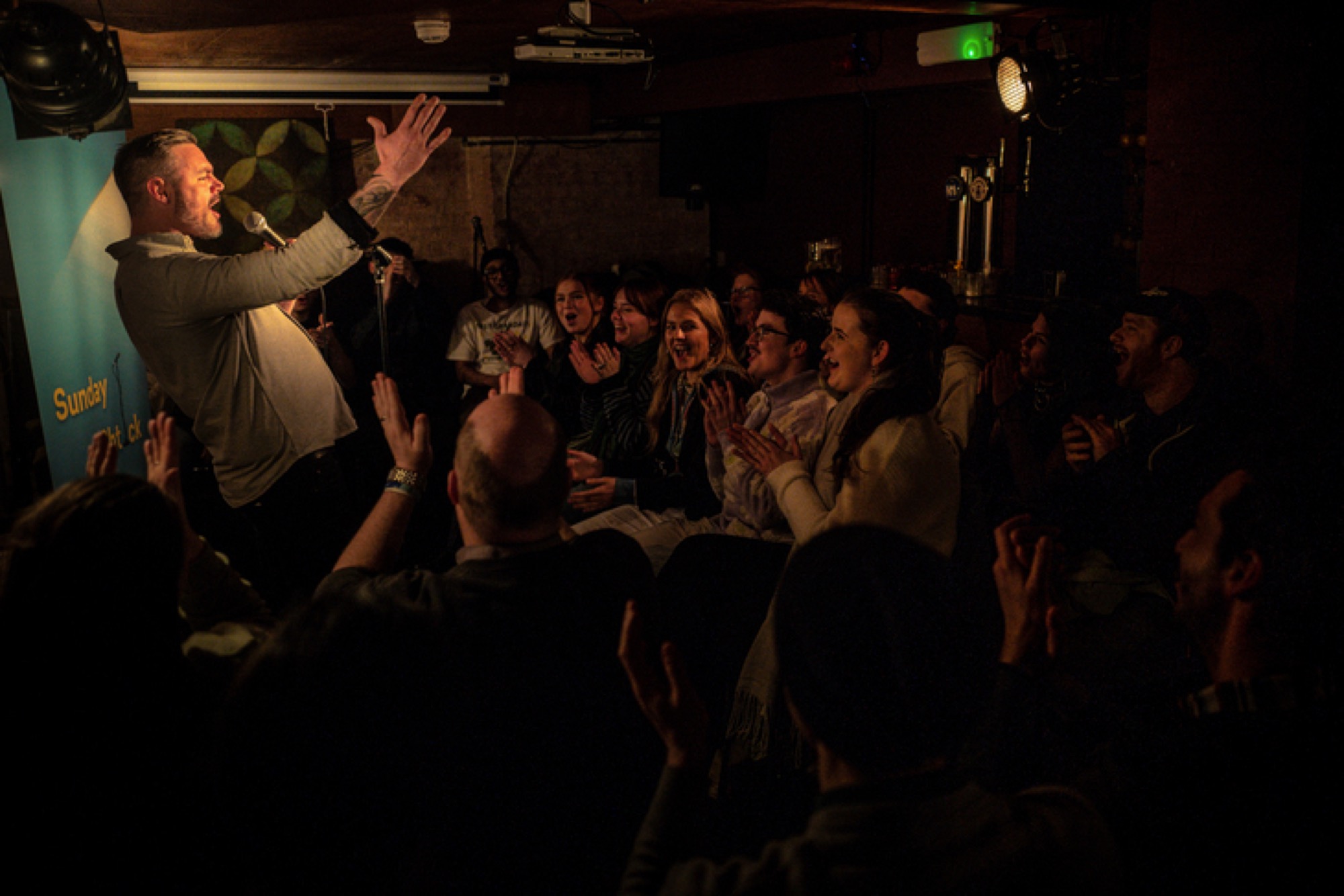 A comic reaching toward a laughing crowd from the stage.